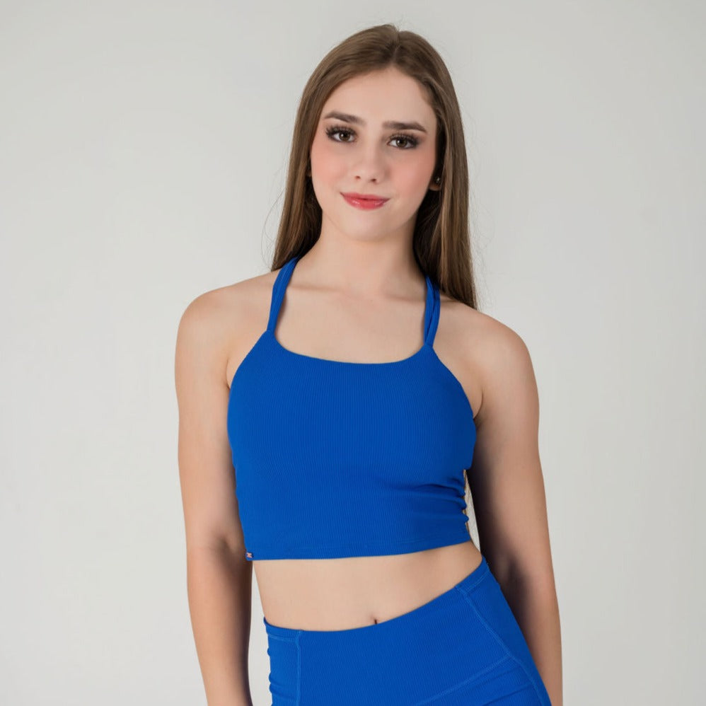 A young woman with long brown hair smiles softly at the camera, wearing the Incanto Ribbed Tank in cobalt blue and matching high-waisted bottoms, standing against a plain light gray background.