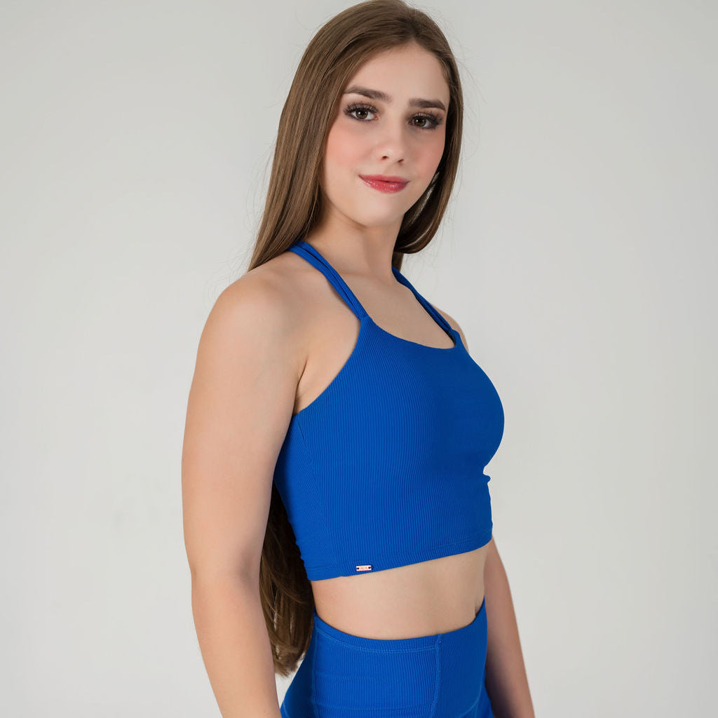 A woman with long brown hair wears the Incanto Ribbed Tank in cobalt blue, paired with matching blue pants, standing against a plain light background and gazing at the camera with a subtle smile.