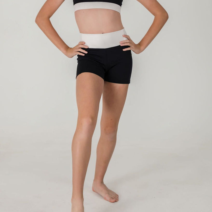 A person stands barefoot on a white background, wearing Chic Shorts - Charcoal (FINAL SALE) and a black and white sports top, with hands on their hips. Their face is not shown.