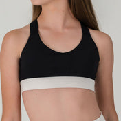 A person with long brown hair models the Chic Top - Charcoal-FINAL SALE, a black color-block sports bra with a wide light gray band, standing slightly turned against a plain background.