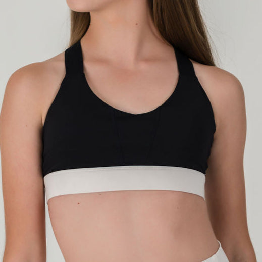 A person with long brown hair models the Chic Top - Charcoal-FINAL SALE, a black color-block sports bra with a wide light gray band, standing slightly turned against a plain background.