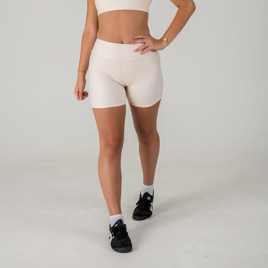 A woman stands against a plain background wearing Airsoft Shorts - Champagne with a matching sports bra, gold wristwatch, black sneakers, and white socks. Her left hand rests on her hip, and her face is not visible.