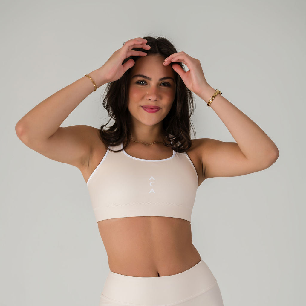 A woman in a Champagne Airsoft Sports Top stands against a plain background, smiling with her hands raised. The moisture-wicking fabric keeps her comfortable and provides maximum support.