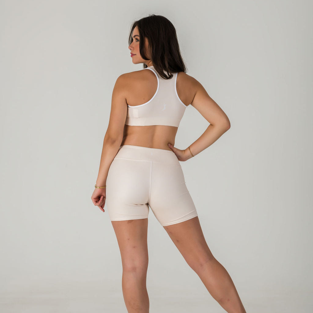 A woman faces away, wearing Airsoft Shorts in Champagne and a matching beige sports bra, both crafted from soft stretch fabric. She poses with one hand on her hip against a plain light gray background.