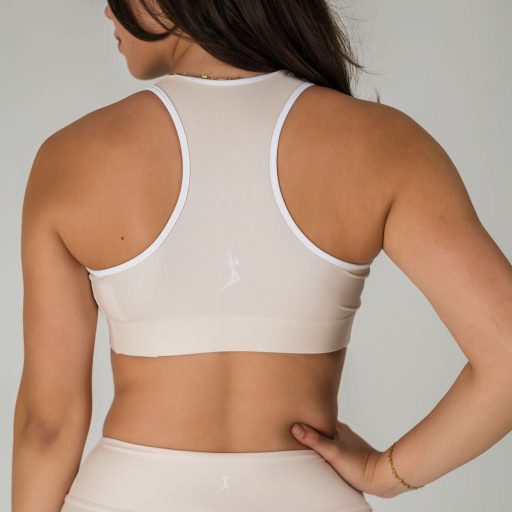 A woman with long brown hair and a gold bracelet is seen from the back wearing the Airsoft Sports Top - Champagne and matching leggings, featuring white trim, a small white logo, and moisture-wicking fabric for maximum support.