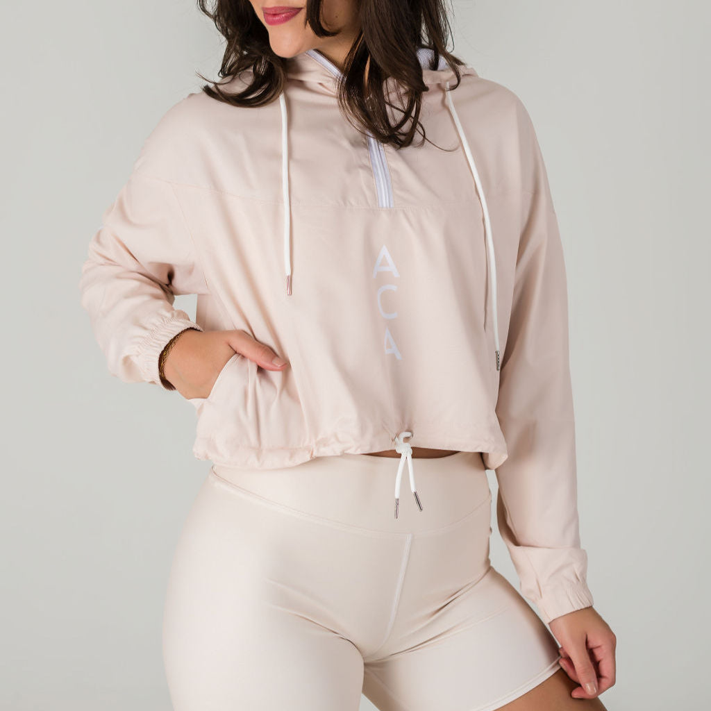 A woman models the Airsoft Anorak - Champagne, a light beige hooded pullover with ACA printed vertically and breathable mesh lining, paired with matching high-waisted bicycle shorts, posing confidently against a plain background.