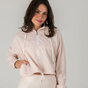 A woman with medium-length brown hair smiles in the Airsoft Anorak - Champagne, a light pink cropped hoodie with a half-zip, ACA letters on the front, and breathable mesh lining. She pairs it with matching leggings against a plain background.