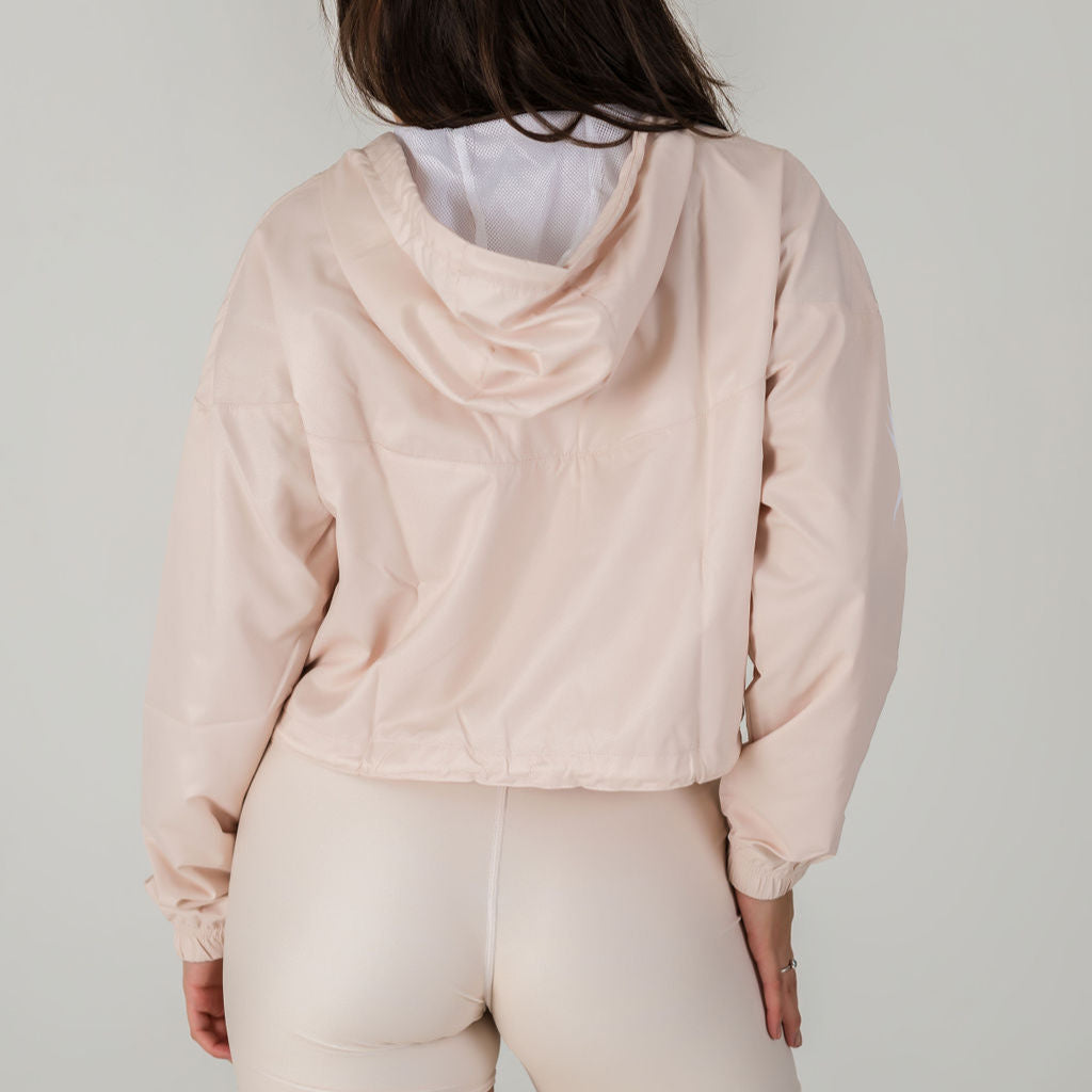 A person with long brown hair is shown from the back wearing the Airsoft Anorak in Champagne—a light beige, water-resistant jacket with a breathable mesh lining—and matching high-waisted leggings against a plain, light background.