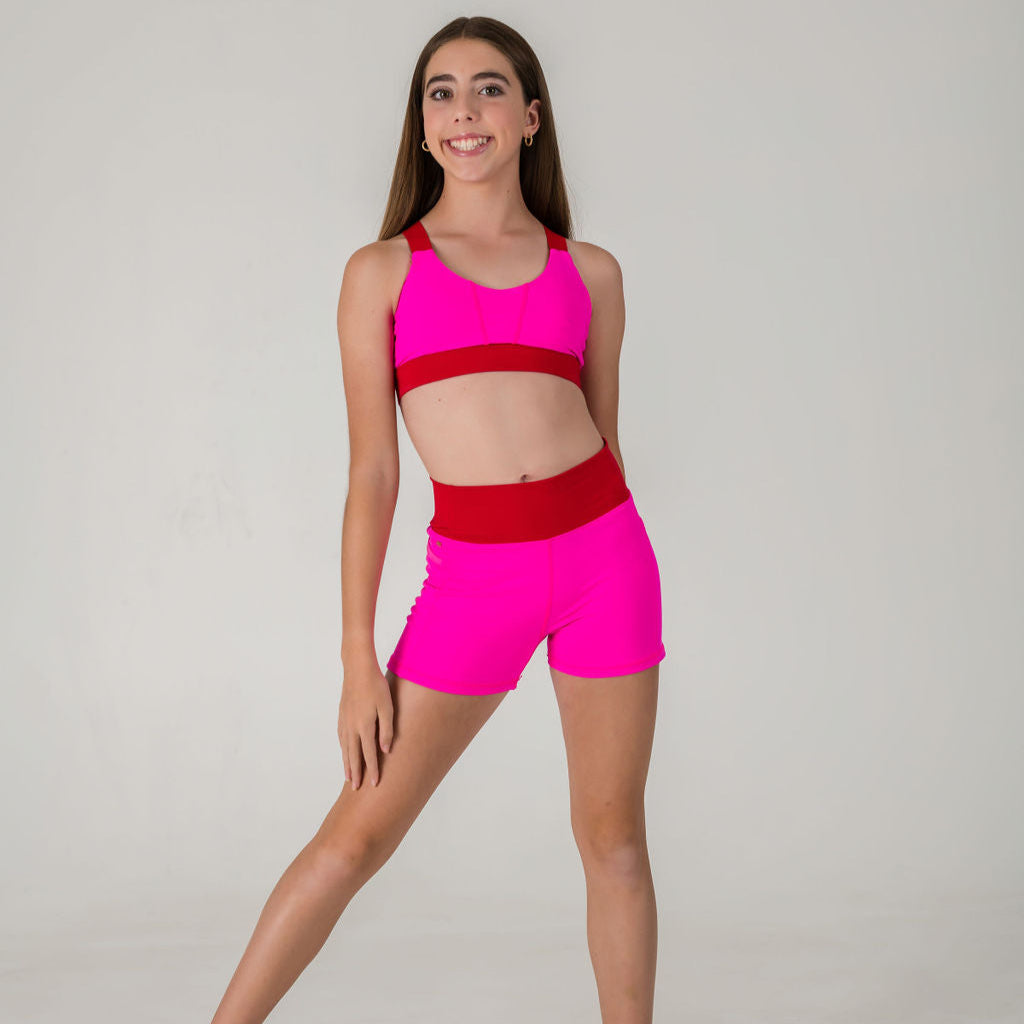 A young girl poses and smiles in the Chic Shorts - Punch Pink - FINAL SALE, paired with a matching pink sports bra featuring a red band, set against a plain light background.
