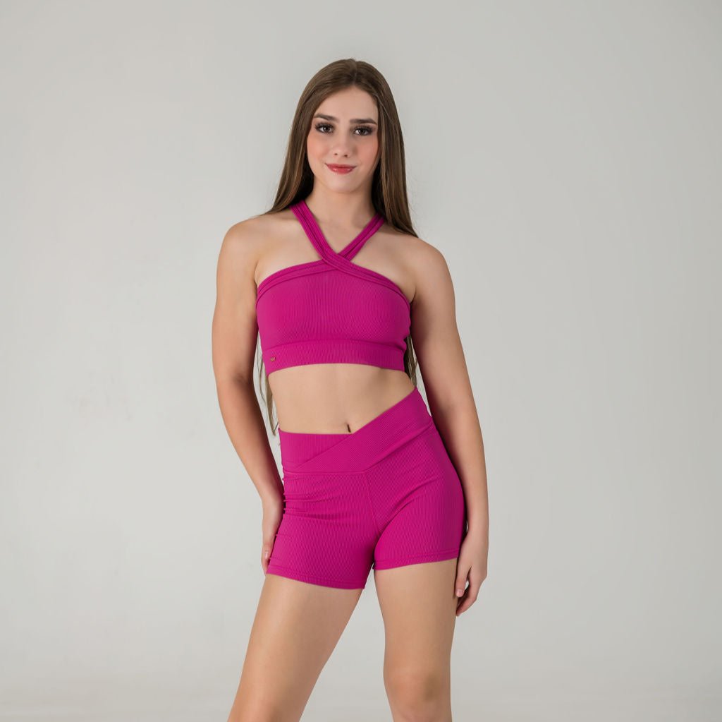 A young woman with long brown hair smiles at the camera, wearing the Iconic Shorts - Pink and a matching crop top, standing against a plain light background with one hand resting on her thigh.