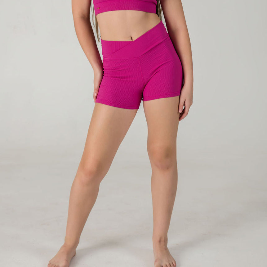 A person stands in front of a plain light background, wearing Iconic Shorts - Pink and a matching magenta sports bra. Their head is out of frame, and their arms are relaxed at their sides.