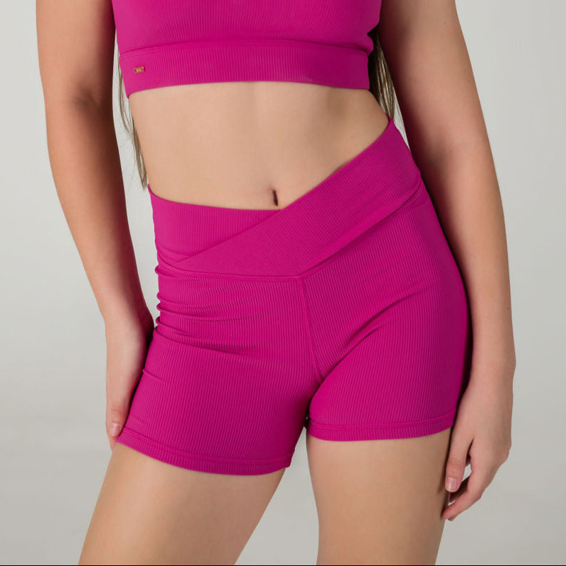 Someone models the Iconic Shorts - Pink, featuring ribbed fabric and a V-front waist, paired with a matching bright pink crop top against a plain light background. Their face is not visible.