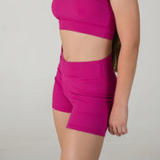 A woman models the Iconic Shorts - Pink with a fuchsia sports bra, standing against a plain light background. Her face is not shown.