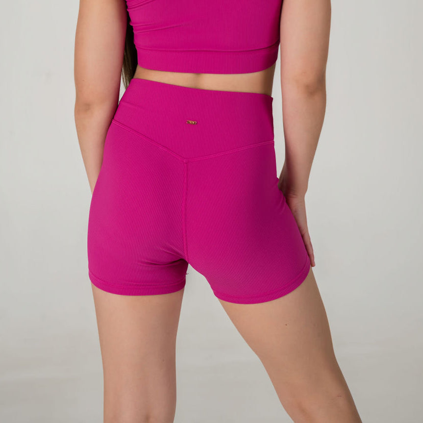 A person stands facing away from the camera, wearing a bright pink crop top paired with the Iconic Shorts - Pink. The background is plain and light-colored.