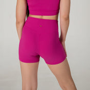 A person stands facing away from the camera, wearing a bright pink crop top paired with the Iconic Shorts - Pink. The background is plain and light-colored.