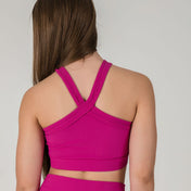 A woman with long brown hair is seen from the back, wearing the Iconic Top - Pink featuring ribbed fabric and crisscross straps, paired with matching high-waisted bottoms against a plain light background.