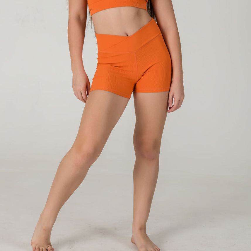 A woman stands barefoot in a studio against a light background, wearing Iconic Shorts - Orange and a matching ribbed sports bra. Her arms rest by her sides, and her long hair falls over one shoulder.
