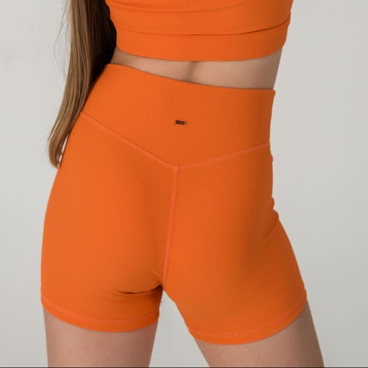 A person stands against a plain light background, shown from the back, wearing Iconic Shorts - Orange and a matching orange ribbed fabric sports bra.