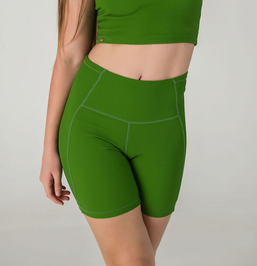 A woman models Incanto Ribbed Shorts in forest green with a matching ribbed crop top. She stands before a plain light gray background, her head out of frame and right hand resting by her side.