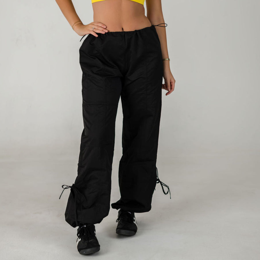 Wearing WOW Parachute Pants in black, paired with black sneakers and a cropped yellow top, a person stands against a plain light background, hands on hips and face out of frame.