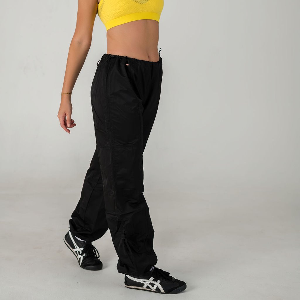 A person wearing WOW Parachute Pants in black with an adjustable waist, a yellow crop top, and black athletic shoes walks against a plain light background. Their head is not visible in the frame.