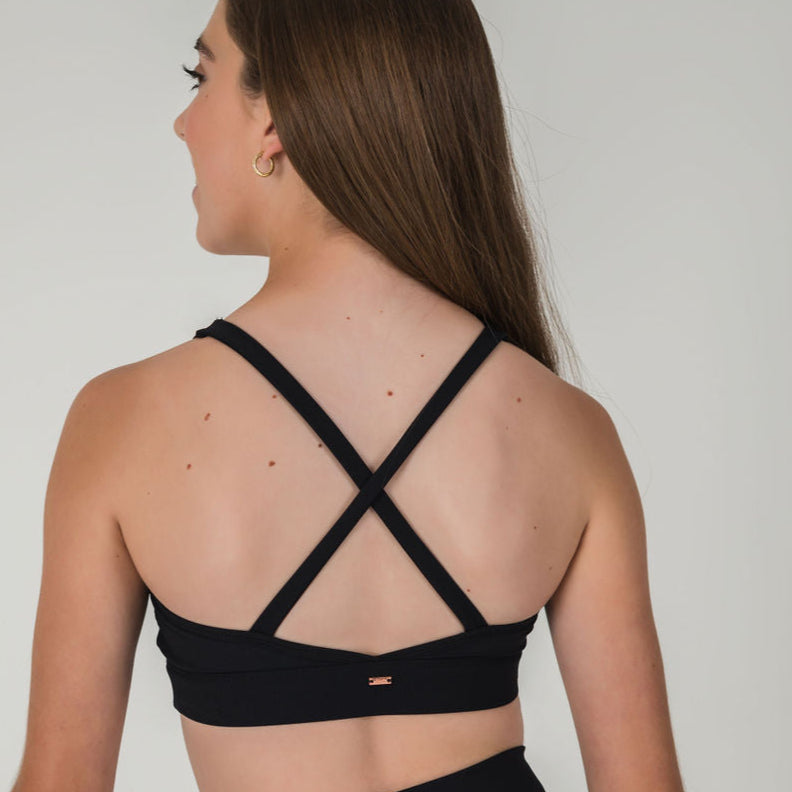 A young woman with long brown hair is seen from behind wearing the 2Iconic Sport Bra, featuring crisscross straps. She has light skin and gold hoop earrings, adding style to her athletic look against a plain light gray background.