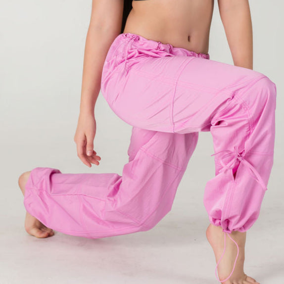 A person wearing WOW Parachute Pants in a loose fit pink style kneels on one knee against a plain light background, with only the pants visible as their upper body and face are not shown.