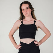 A young woman with long brown hair stands against a light background, serious-faced, wearing the black ICONIC2 Tank with removable pads and high-waisted leggings. Her hands rest on her hips.