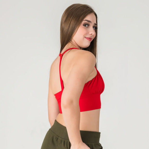 A young woman with long brown hair poses sideways and smiles against a plain light gray background, wearing the XOXO Top - Cherry Red with a ruched front and olive green pants.