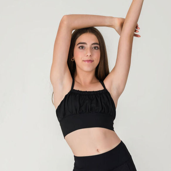 A young woman stretches her arms above her head while wearing the XOXO Top in Midnight Black, standing against a plain light background.