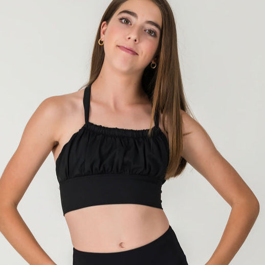 A young woman with straight brown hair wears a sleeveless XOXO Top - Midnight Black (FINAL SALE) featuring a ruched front, posing with her hands on her hips against a plain light background.