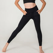 A person in a black crop top and XOXO Skinny Joggers - FINAL SALE with pockets stands barefoot against a white background, posing with legs apart and hands on hips.