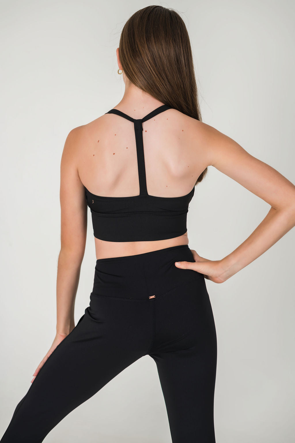 A woman with long brown hair stands against a light background, hand on hip, wearing XOXO Skinny Joggers - FINAL SALE and a black sports bra with T-shaped straps.
