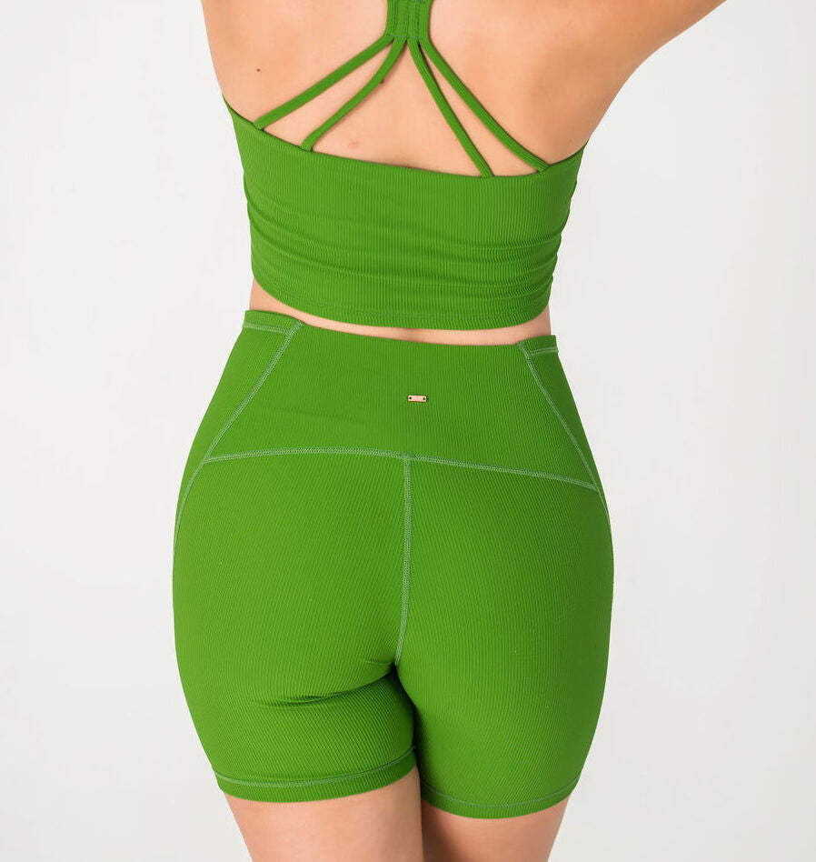 A person is seen from the back wearing the Incanto Ribbed Shorts - Forest Green, a high-waisted style made from ribbed fabric, paired with a matching green crop top with criss-cross straps. The background is plain and light-colored.