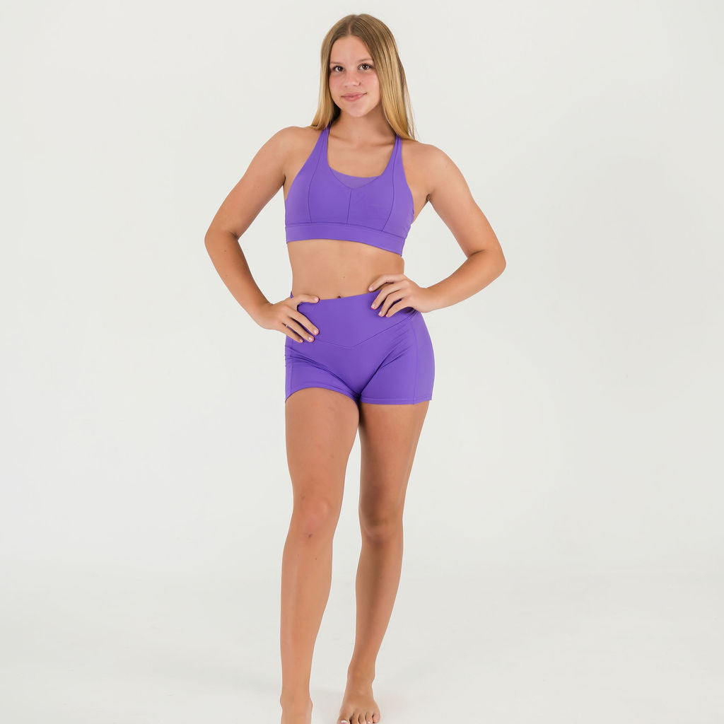 A young woman, barefoot against a white background, wears the Bonbon Royal Purple Top with adjustable crisscross straps and matching shorts, her right hand on her hip and left arm relaxed as she smiles confidently.