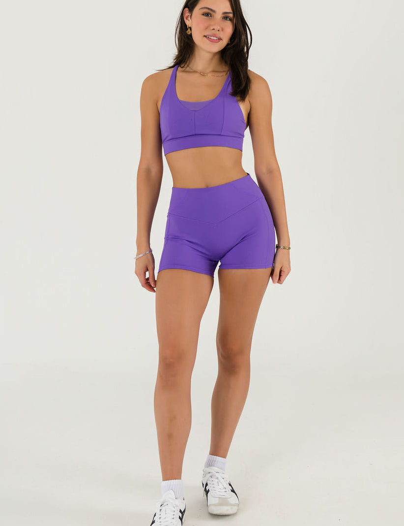 A person models the Bonbon Shorts in Royal Purple with a matching sports bra and white sneakers against a plain white background. Their head is not visible in the image.