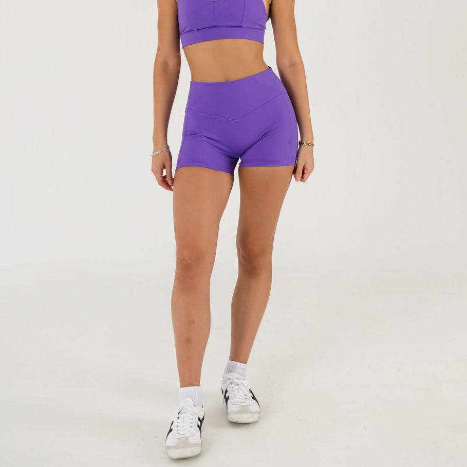 A person models the Bonbon Shorts in Royal Purple with a matching sports bra and white sneakers against a plain white background. Their head is not visible in the image.