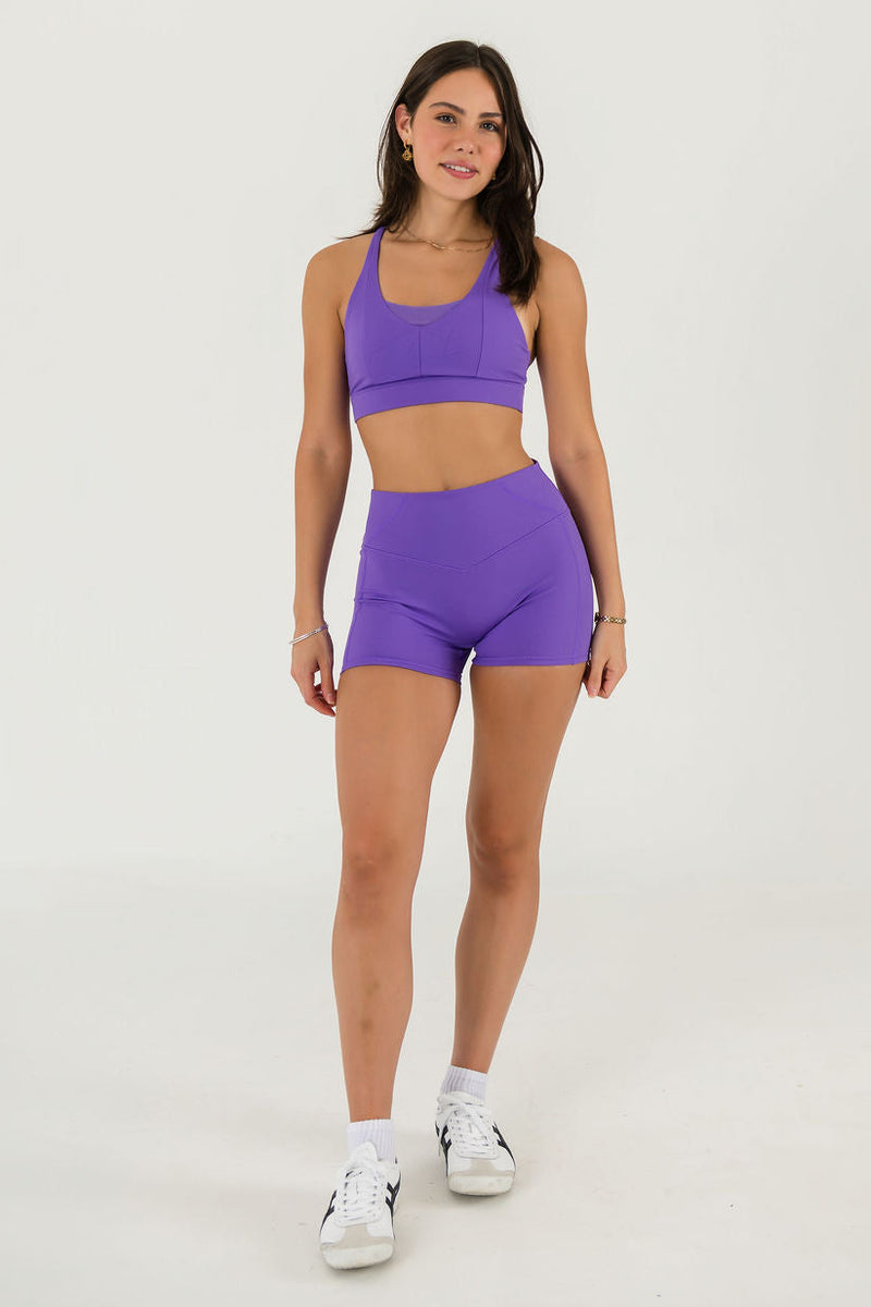 A person models the Bonbon Shorts in Royal Purple with a matching sports bra and white sneakers against a plain white background. Their head is not visible in the image.