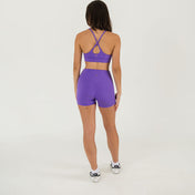 A woman faces away from the camera wearing Bonbon Royal Purple Shorts, a matching sports bra, and white sneakers against a plain white background.