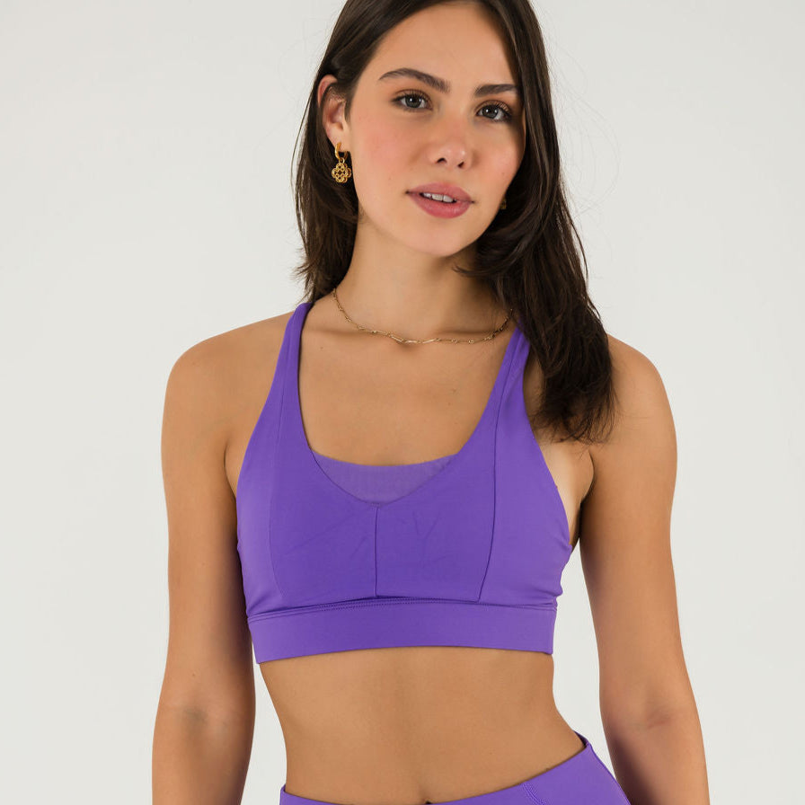 A woman with long brown hair and minimal jewelry stands facing forward in Bonbon Royal Purple Shorts and a matching sports bra, her expression neutral against a plain light background.