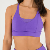 A woman in Bonbon Royal Purple Shorts and a matching sports bra stands against a plain white background, smiling slightly.