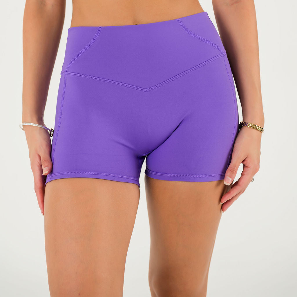 A person stands against a plain white background wearing Bonbon Shorts - Royal Purple, featuring a high waist, seamless front, and comfortable elastic fit; their hands rest by their sides with simple bracelets on each wrist.