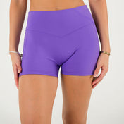 Wearing Bonbon Royal Purple Shorts, a person stands against a plain white background. Only their lower torso and upper legs are visible, with bracelets adorning both wrists.