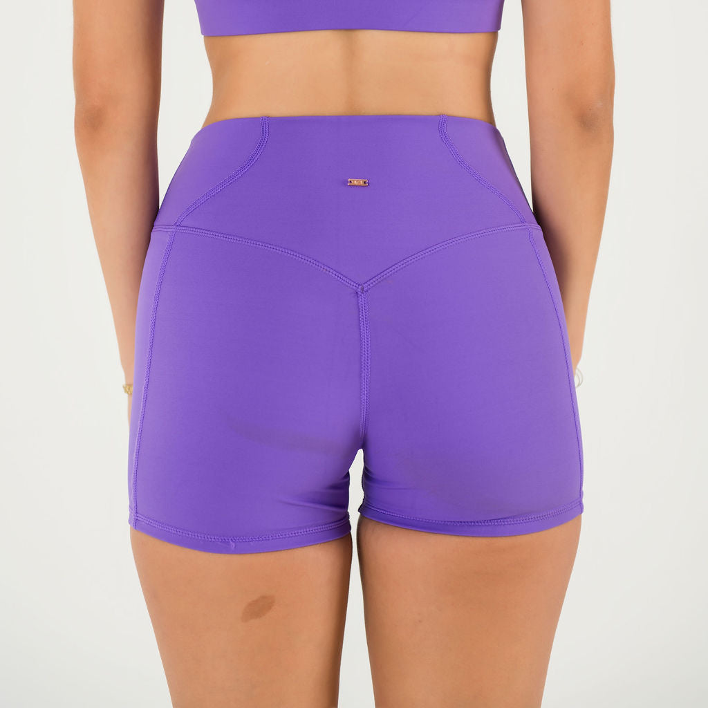A woman stands with her back to the camera against a plain white background, wearing Bonbon Royal Purple Shorts and a matching sports bra.