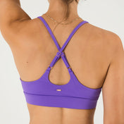 A woman with her hair tied up, wearing a gold chain necklace and the Bonbon Royal Purple Top with a scoop neckline and adjustable crisscross back straps, stands facing away from the camera against a plain white background.