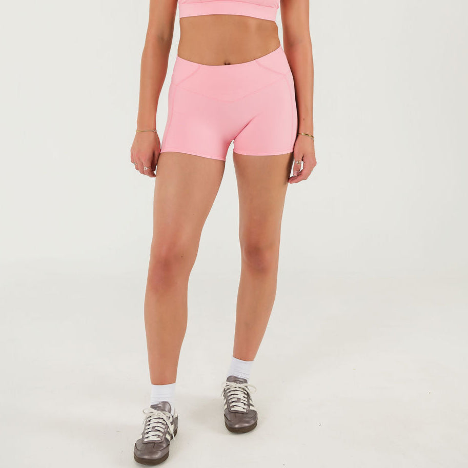 A person models Bonbon Vivid Pink Shorts made from soft, stretchy fabric, paired with a pink sports bra. Their head isnt visible as they stand against a plain white background, also wearing white socks and gray sneakers.