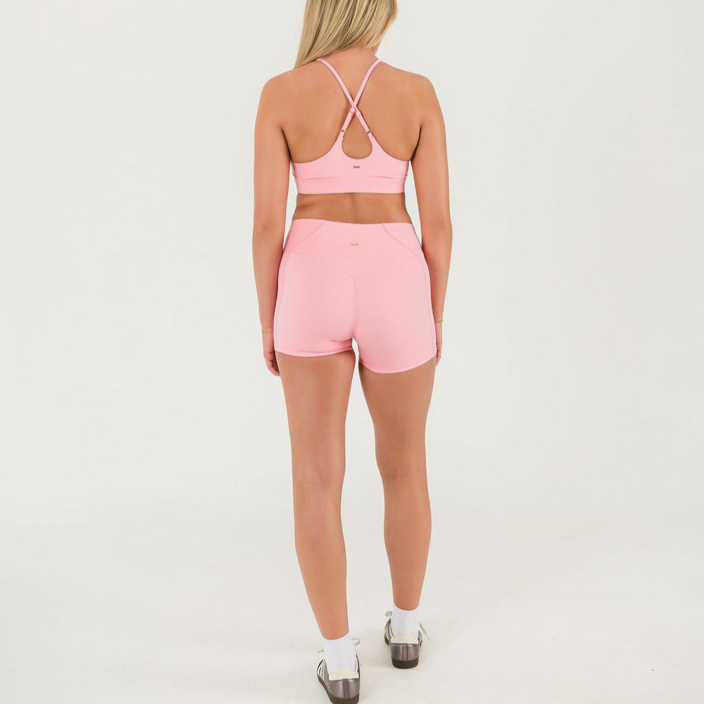 A woman stands facing away from the camera wearing a light pink sports bra and Bonbon Vivid Pink Shorts made from soft, stretchy fabric, paired with white socks and sneakers against a plain white background.