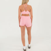 A woman stands facing away from the camera wearing a light pink sports bra and Bonbon Vivid Pink Shorts made from soft, stretchy fabric, paired with white socks and sneakers against a plain white background.