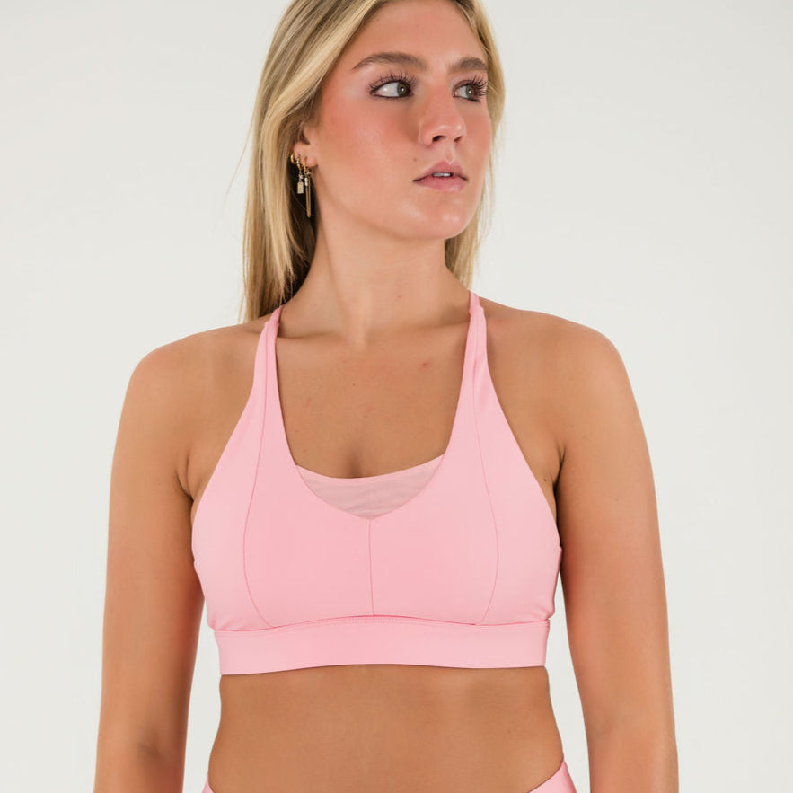 A woman with long blonde hair wears the Bonbon Top - Vivid Pink, a scoop neckline sports bra with matching bottoms, standing against a plain white background and looking slightly to her left.