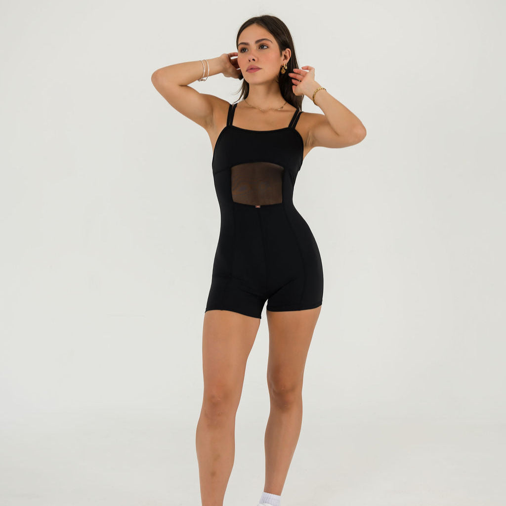 A woman poses against a white background wearing the Gia Unitard, a black fitted piece with mesh torso panels. Her hands are raised near her head as she looks slightly to the side.